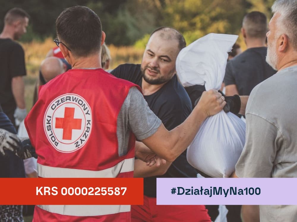 Mężczyzna w kamizelce Polskiego Czerwonego Krzyża podaje worek z piaskiem w czasie akcji stawiania wałów przeciwpowodziowych
